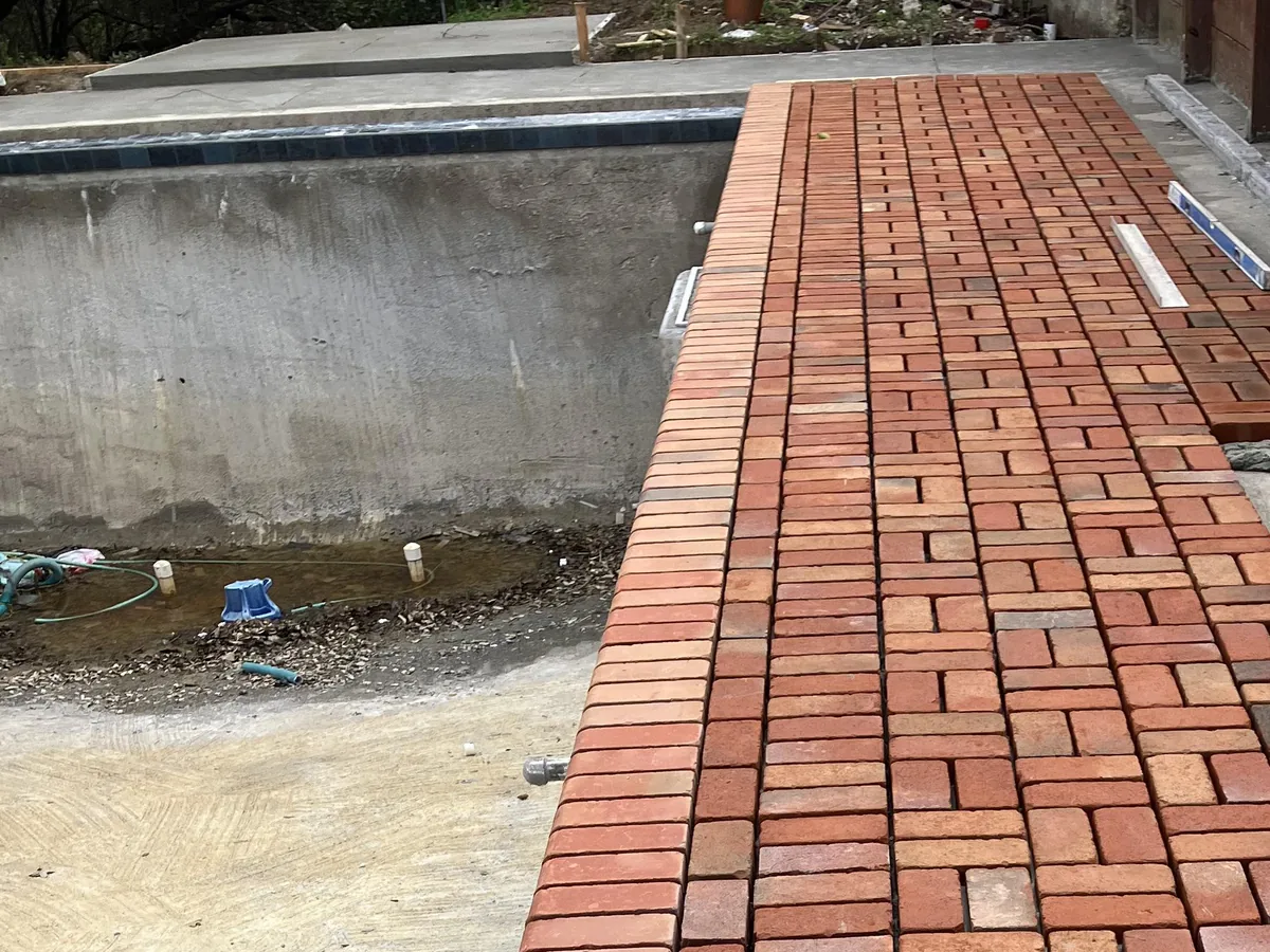 Reclaimed clay brick pavers going down in a basketweave pattern with a soldier border on the concrete deck beside a new pool, brick pavers