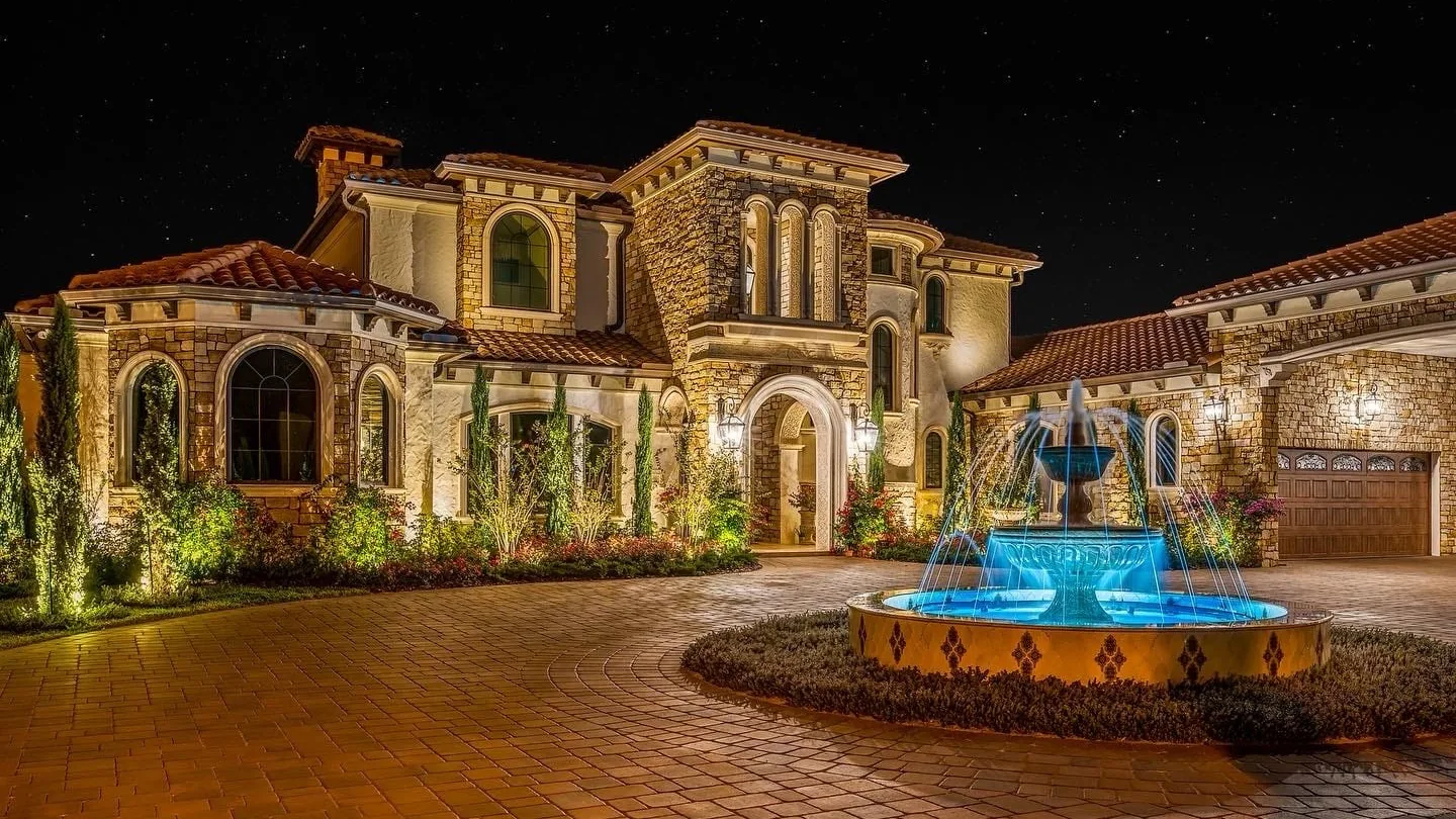 Mediterranean-style home faced in natural stone with arched windows and an arched stone entry, lit at night beside a tiered fountain, stone masonry