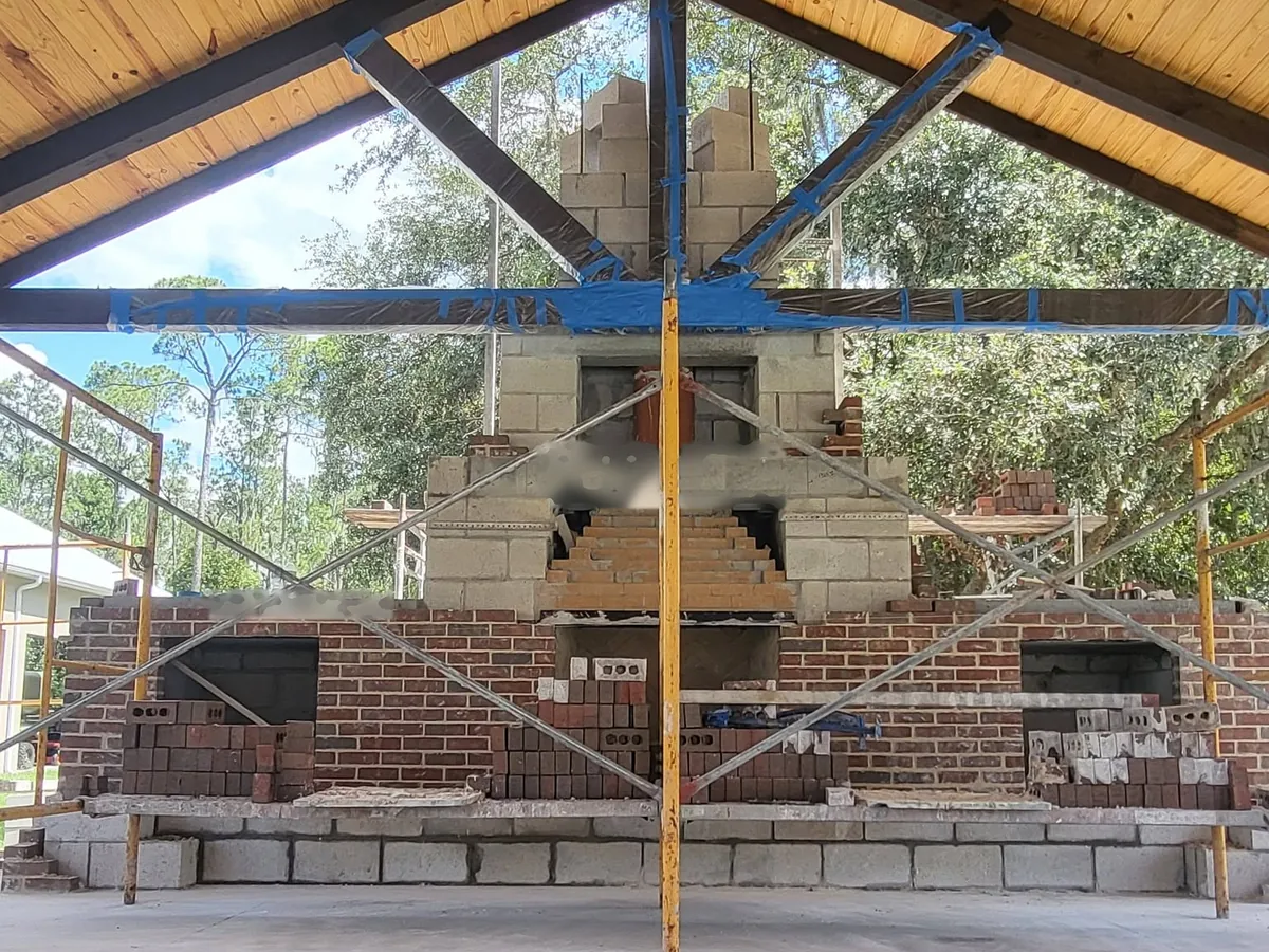 Brick outdoor fireplace wall with two side niches and a concrete block chimney core rising through a timber pavilion roof, with scaffolding, outdoor fireplaces