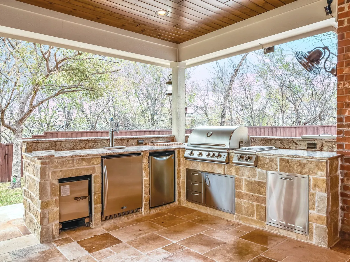 L-shaped natural stone outdoor kitchen with a built-in grill, stainless doors and a sink under a covered patio, outdoor kitchens