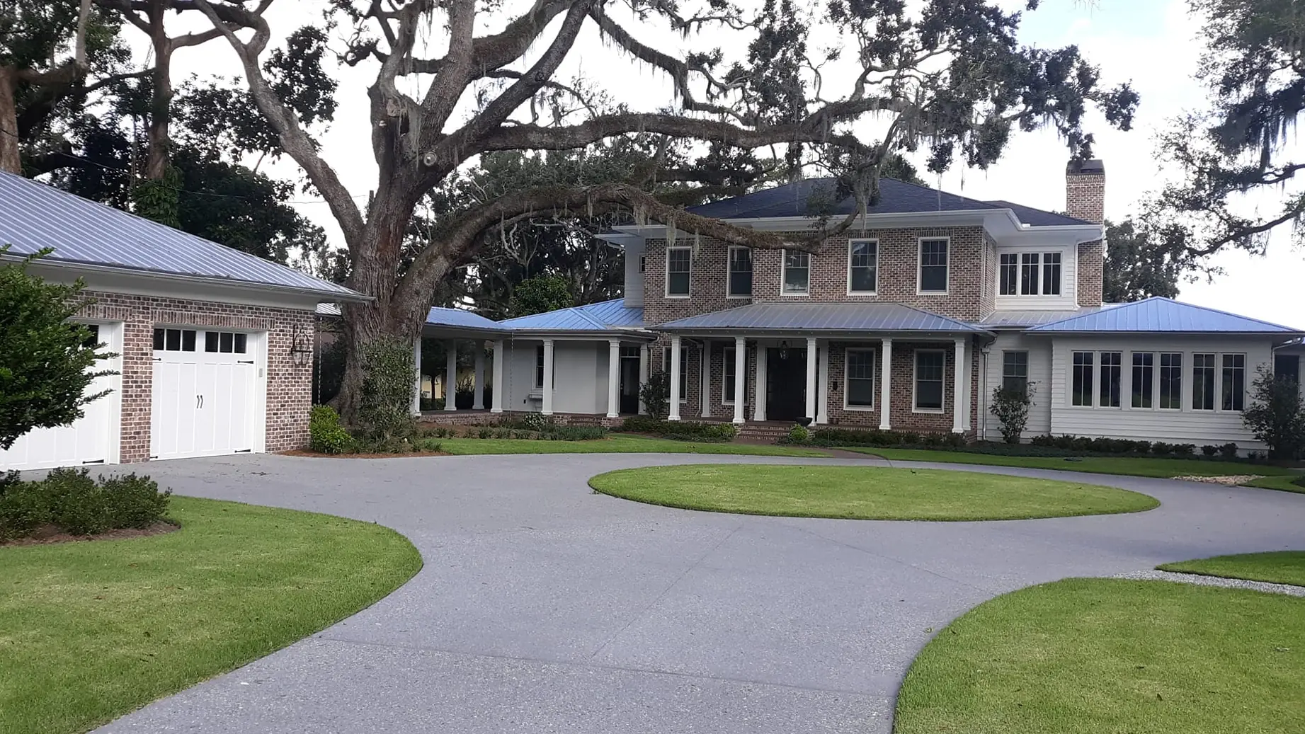 Two-story brick house with a wraparound porch and a curved charcoal concrete driveway under a live oak, masonry