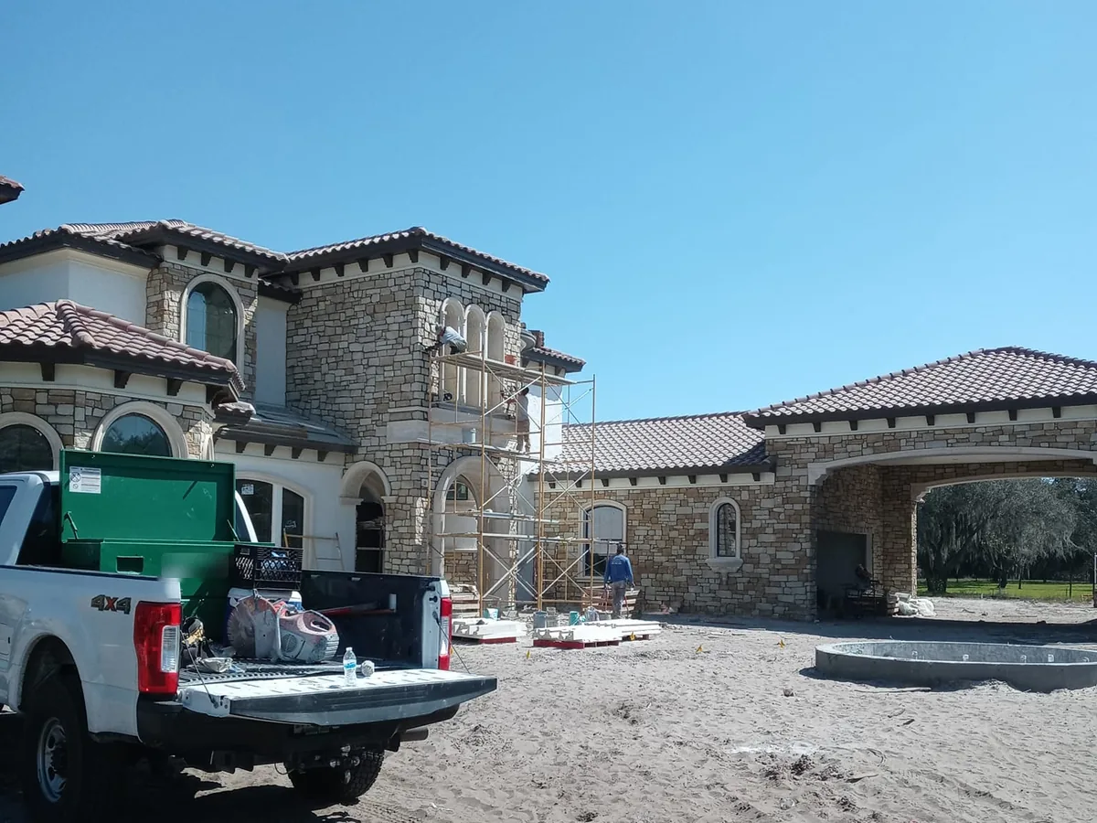 Large house faced in natural stone during construction, with scaffolding at an arched tower window, stone masonry